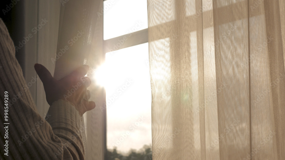 Naklejka premium Close up woman hand opening transparent curtain tulle by window to enjoy early morning sunrise at home. Have fun warm energy sun shine sunlight at home and thinking dreaming looking outside at street