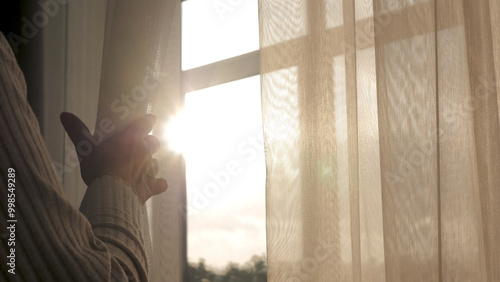 Close up woman hand opening transparent curtain tulle by window to enjoy early morning sunrise at home. Have fun warm energy sun shine sunlight at home and thinking dreaming looking outside at street