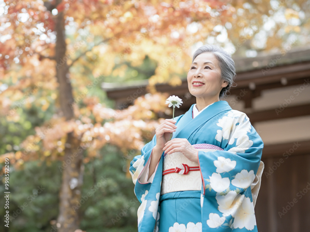 Elderly Japanese woman in blue floral kimono holding a flower, smiling in a beautiful autumn garden Elderly Japanese woman in blue floral kimono holding a flower, smiling in a beautiful autumn garden
