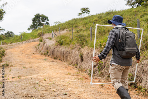 Colombian Latino biologist carrying quadrat frames on his back while walking along a path, preparing to measure pasture growth