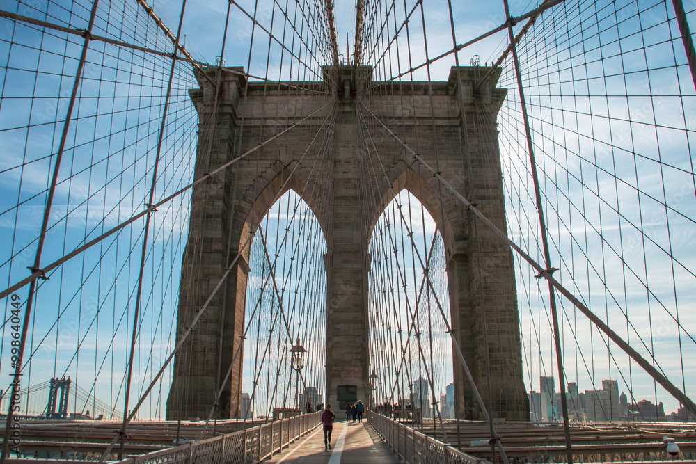 Fototapeta premium Personas cruzando el puente de Brooklyn en la zona peatonal. Paisaje urbano en una fría mañana de otoño. Al fondo, los edificios de Brooklyn a orillas del río Este. Nueva York, Estados Unidos. 2019.