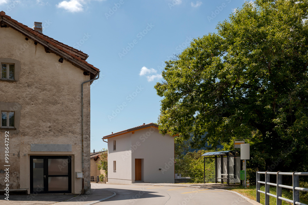 Exterior of the newly renovated house, with the road running past the wall, on a beautiful sunny day with blue skies. To the left is an old village house and across the street is the bus stop, no one Exterior of the newly renovated house, with the road running past the wall, on a beautiful sunny day with blue skies. To the left is an old village house and across the street is the bus stop, no one