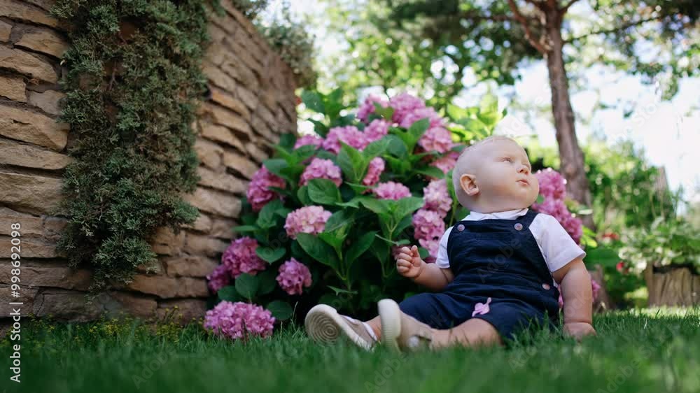 Little lovely kid wearing blue romper sits in the grass. Low angle view at the blond baby boy sitting in nature. Little lovely kid wearing blue romper sits in the grass. Low angle view at the blond baby boy sitting in nature.