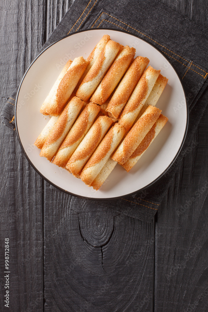 Russian cigarettes are thin delicate wafers rolled into a roll and baked until golden brown and crispy close-up on a plate on the table. Vertical top view from above Russian cigarettes are thin delicate wafers rolled into a roll and baked until golden brown and crispy close-up on a plate on the table. Vertical top view from above