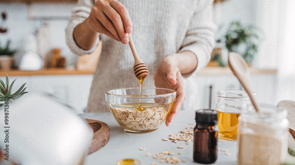 A woman is standing in a kitchen, smiling as she pours honey from a wooden spoon into a glass bowl filled with oatmeal. The scene is set against a blurred background. A woman is standing in a kitchen, smiling as she pours honey from a wooden spoon into a glass bowl filled with oatmeal. The scene is set against a blurred background.