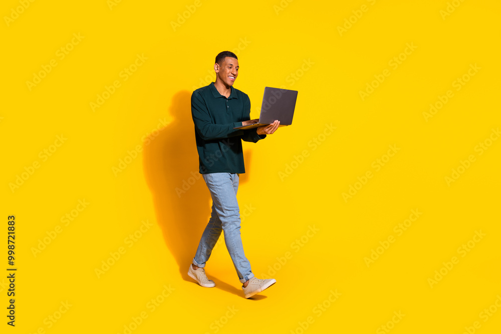 Full size photo of handsome young guy walking hold netbook dressed stylish green garment isolated on yellow color background Full size photo of handsome young guy walking hold netbook dressed stylish green garment isolated on yellow color background