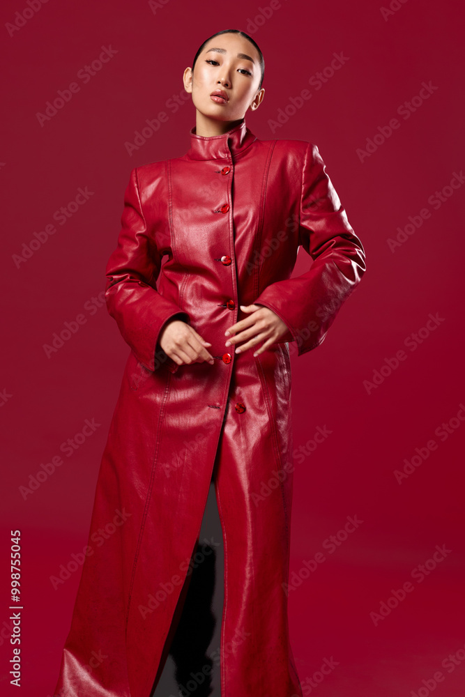 Fashion asian woman in a red leather coat posing against a red background with hands on hips Fashion asian woman in a red leather coat posing against a red background with hands on hips