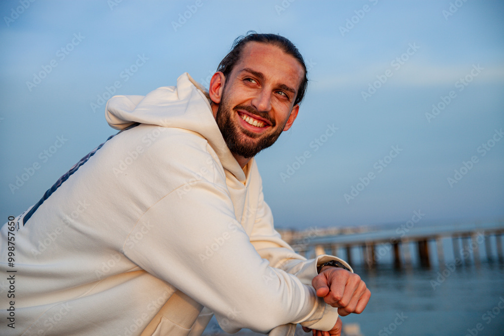 portrait of a handsome and sporty young guy dressed in casual clothes in a seaside location. The model is posing with a smiling expression while standing on a beach at sunset
