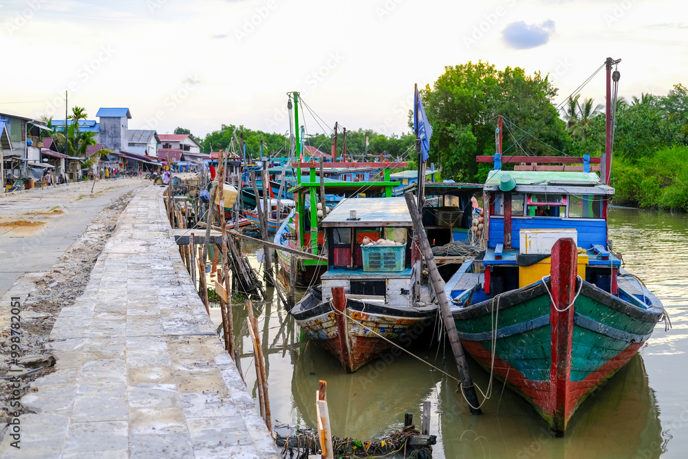 Colorful fishing boats docked along the concrete pier at Pelabuhan Kuala Singkawang, Kalimantan Barat, with village buildings and lush trees in the background, capturing coastal life.