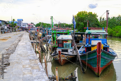 Obraz na płótnie Colorful fishing boats docked along the concrete pier at Pelabuhan Kuala Singkawang, Kalimantan Barat, with village buildings and lush trees in the background, capturing coastal life. w salonie