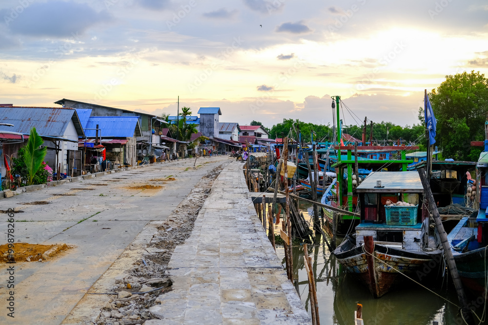Colorful fishing boats docked along the concrete pier at Pelabuhan Kuala Singkawang, Kalimantan Barat, with village buildings and lush trees in the background, capturing coastal life.