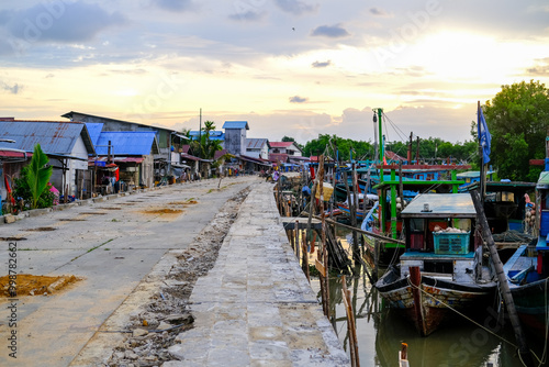 Obraz na płótnie Colorful fishing boats docked along the concrete pier at Pelabuhan Kuala Singkawang, Kalimantan Barat, with village buildings and lush trees in the background, capturing coastal life. w salonie