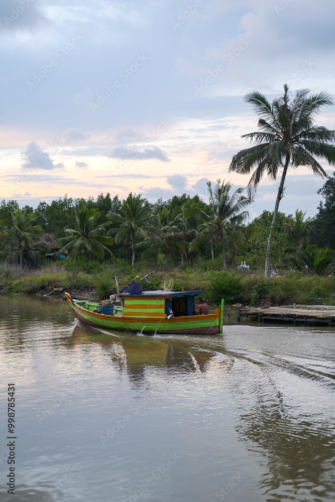 Colorful fishing boats docked along the concrete pier at Pelabuhan Kuala Singkawang, Kalimantan Barat, with village buildings and lush trees in the background, capturing coastal life.