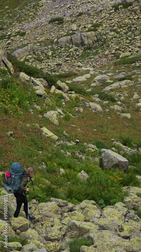 A female backpacker carefully navigates through a rugged mountain landscape filled with large, yellow-lichen-covered boulders and alpine vegetation. Using trekking poles for support, she moves