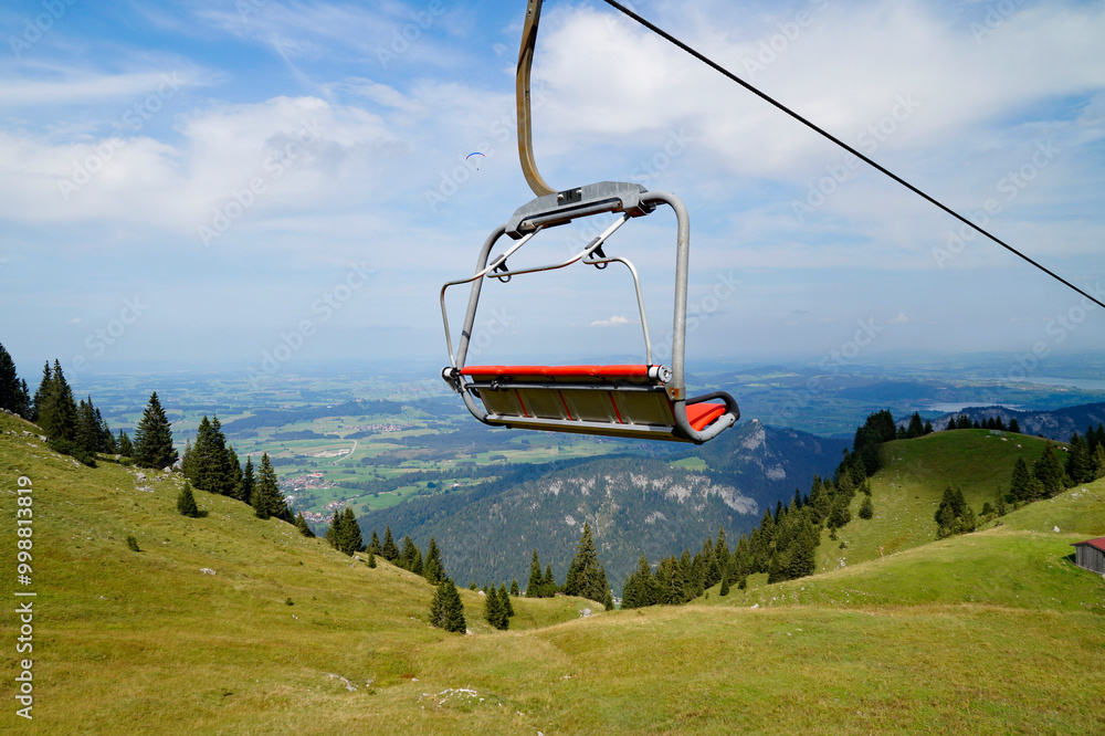 scenic alpine view with a cableway with red chairlift over the Bavarian Alps and lake Forggensee and a paraglider in the background, seen from Breitenberg mountain (Pfronten, Bavaria, Germany) scenic alpine view with a cableway with red chairlift over the Bavarian Alps and lake Forggensee and a paraglider in the background, seen from Breitenberg mountain (Pfronten, Bavaria, Germany)