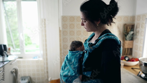 Mother standing in the kitchen with her baby in a carrier, looking preoccupied and thoughtful, portraying a moment of reflection and care in a cozy and nurturing home environment