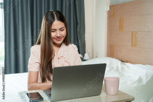 Asian woman working from home sitting at desk in bedroom using laptop wearing pink blouse looking at screen with focus relaxed home office environment cozy working setup.