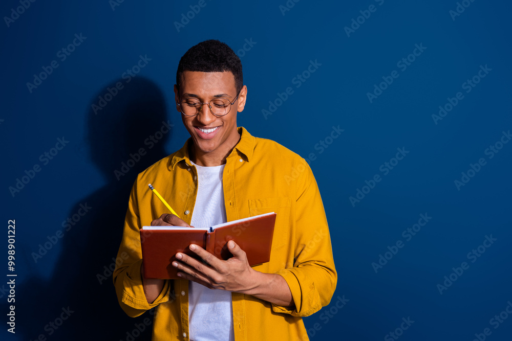 Photo of guy in yellow shirt wearing eyeglasses holding copybook writing details for his project isolated on dark blue color background Photo of guy in yellow shirt wearing eyeglasses holding copybook writing details for his project isolated on dark blue color background