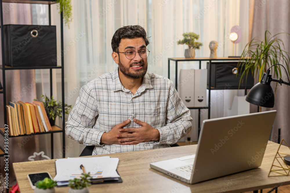 Obraz premium Disappointed Hispanic male freelancer feeling overeating during work process on laptop in home office. Upset Indian businessman freelancer touching stomach and experiencing discomfort and sharp pain.
