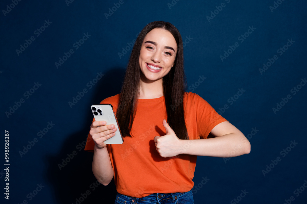 Photo of lovely nice cheerful woman wear orange stylish clothes recommend gadget thumb up isolated on dark blue color background Photo of lovely nice cheerful woman wear orange stylish clothes recommend gadget thumb up isolated on dark blue color background