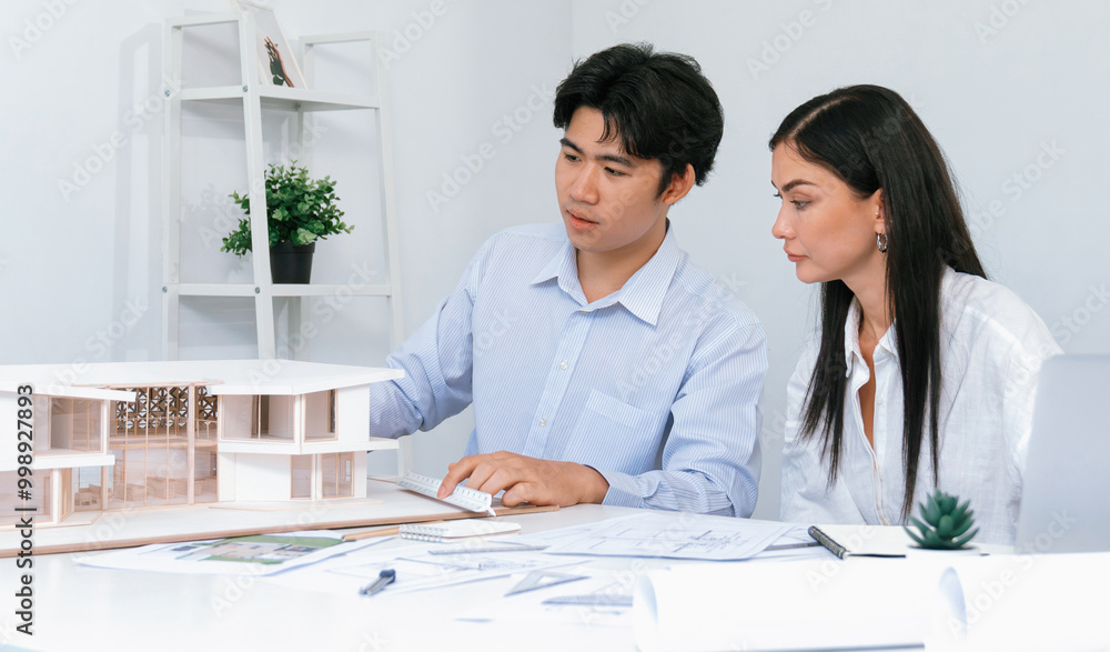 Professional male asian architect using ruler to measure house model length while young beautiful caucasian colleague using laptop to analyzed data on meeting table with house model. Immaculate. Professional male asian architect using ruler to measure house model length while young beautiful caucasian colleague using laptop to analyzed data on meeting table with house model. Immaculate.