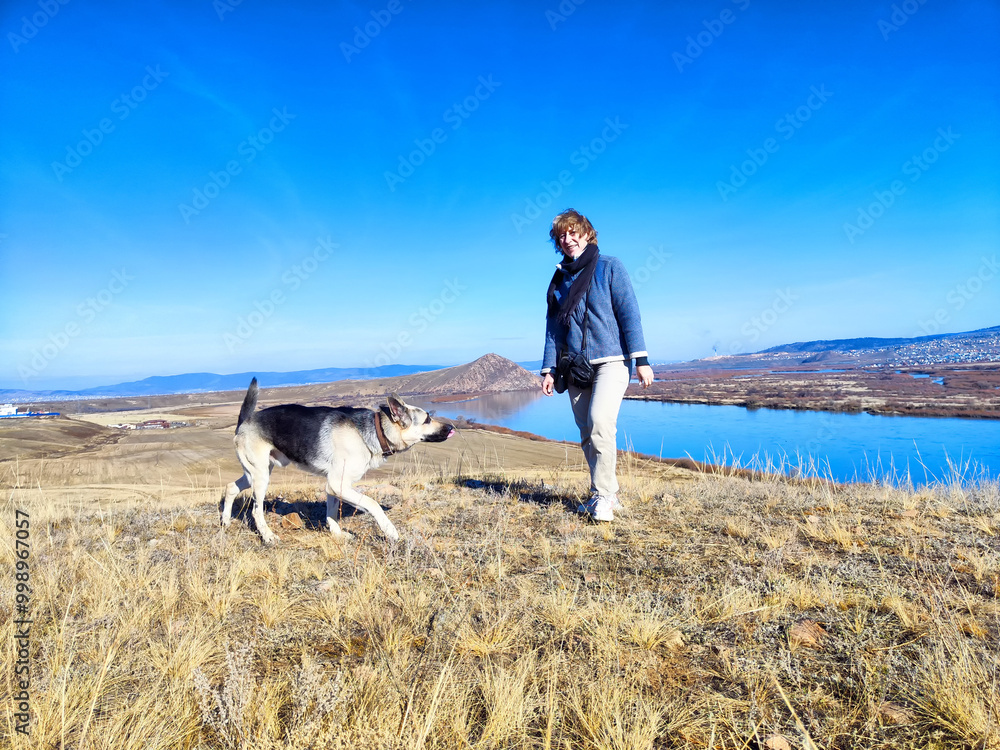 Obraz premium A middle-aged woman with a German Shepherd dog in nature in spring or autumn day with sun and blue sky. A female traveler with a pet on a trip