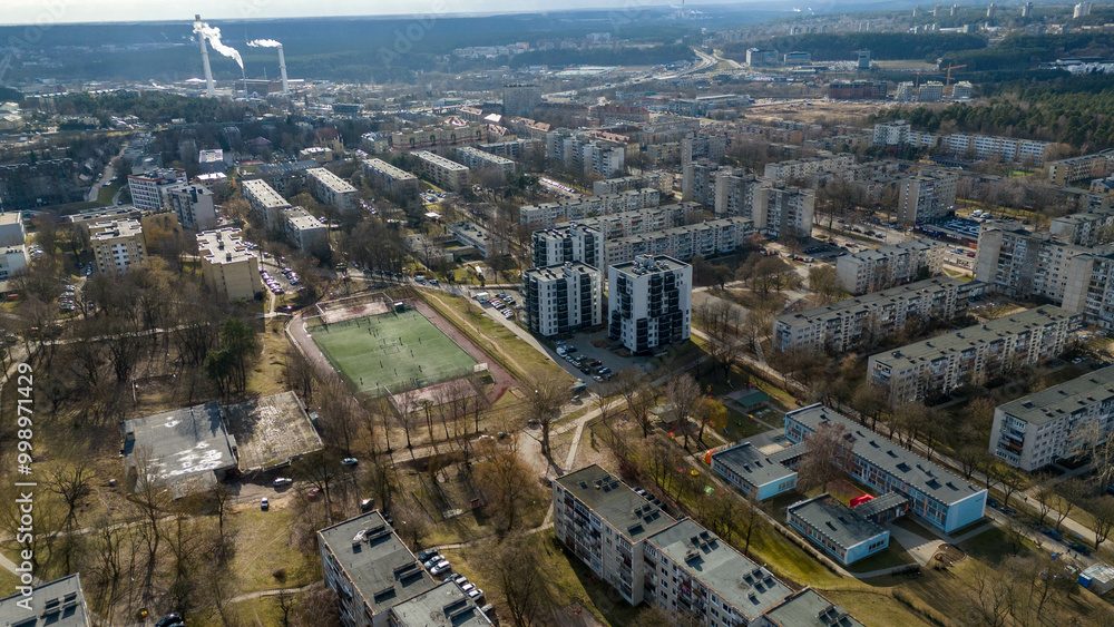 Fototapeta premium Aerial view of a modern urban area featuring a mix of residential and commercial buildings, with green spaces and parking lots. The skyline includes various structures.
