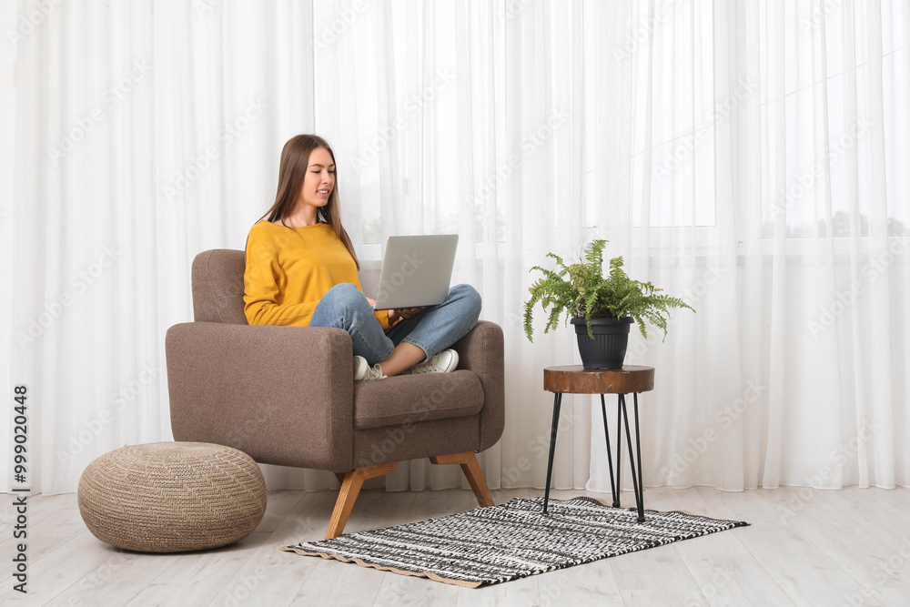 Beautiful young woman with laptop in comfortable armchair near window in living room Beautiful young woman with laptop in comfortable armchair near window in living room