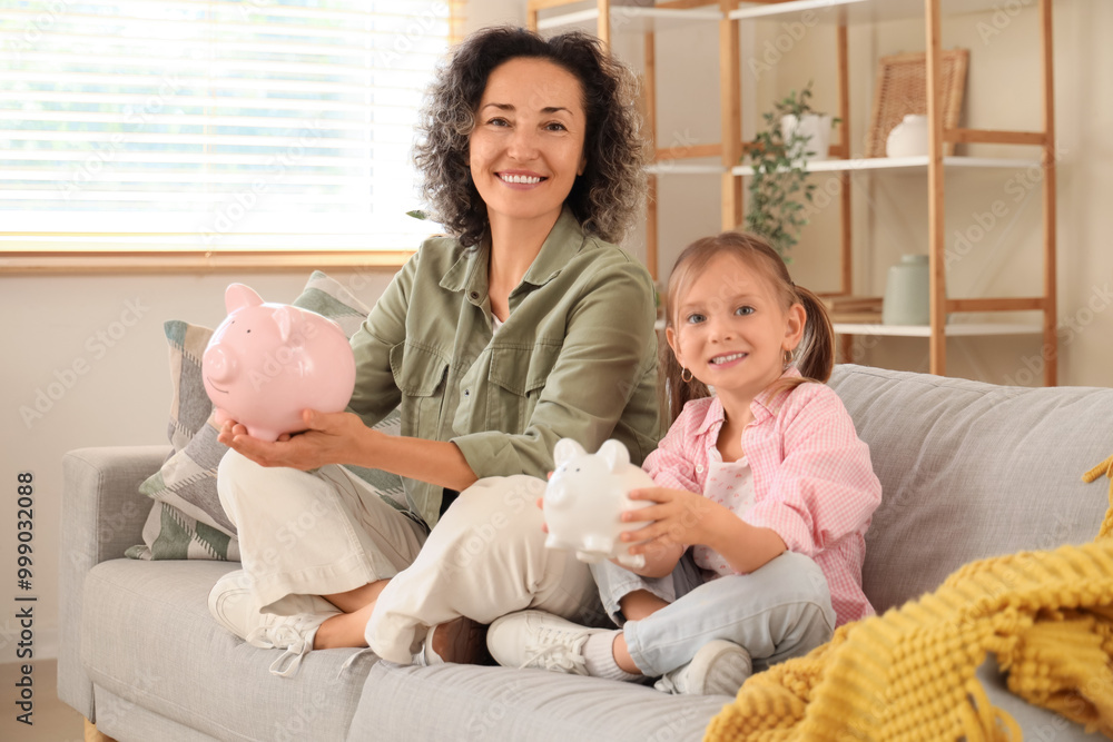 Little girl with her mother and piggy banks sitting on sofa at home Little girl with her mother and piggy banks sitting on sofa at home