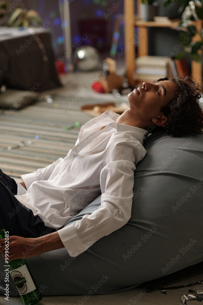 Young asleep man keeping head on edge of bed while sitting on the floor after booze party in messy domestic room Young asleep man keeping head on edge of bed while sitting on the floor after booze party in messy domestic room