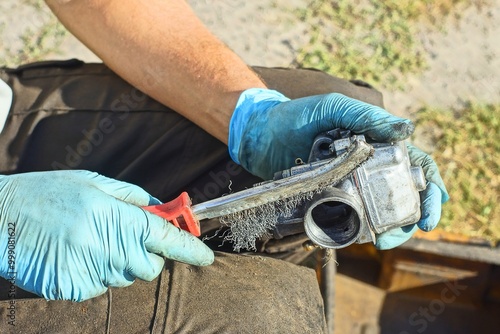 worker with hands in blue rubber gloves sharp with plastic red handle brush for metal cleans industrial gray disassembled iron old carburetor from engine of retro motorcycle in daytime on the street