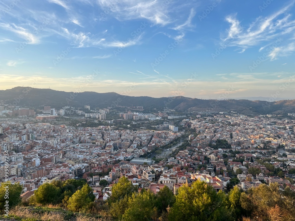 Obraz premium Barcelona Cityscape from Above at Dusk: A panoramic view of Barcelona’s urban sprawl and distant mountains, seen from an elevated vantage point as golden hour light softens the city and sky.