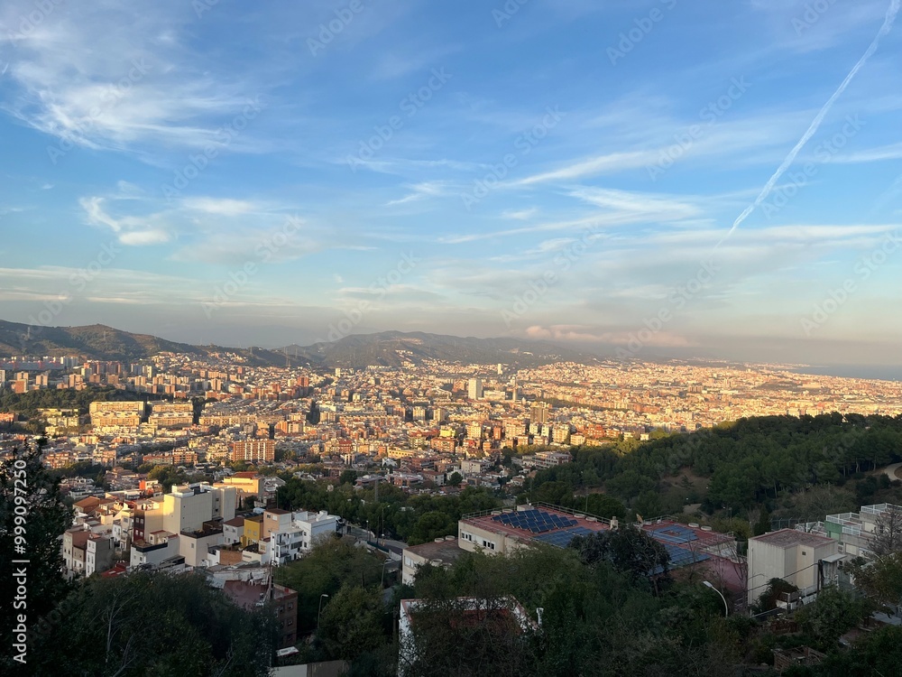 Obraz premium Barcelona Cityscape from Above at Dusk: A panoramic view of Barcelona’s urban sprawl and distant mountains, seen from an elevated vantage point as golden hour light softens the city and sky.