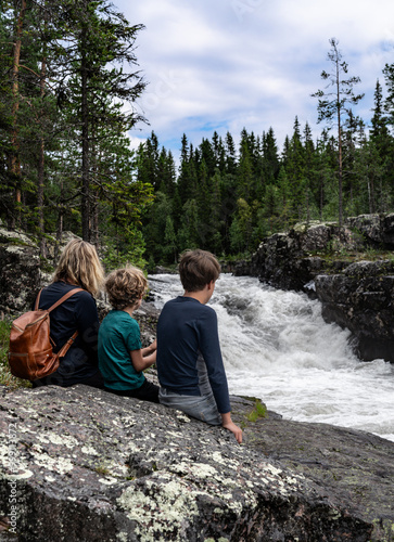 A mother and her two sons sit on a rock, captivated by the powerful Sognstupet Waterfall in Idre Dalarna surrounded by green trees and rushing water in the wilderness of Sweden photo