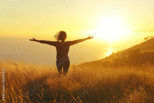 Woman with arms outstretched on a grassy hill, basking in the warm golden light of a beautiful sunrise over the ocean, embodying happiness and peace.