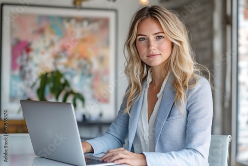 woman working on laptop in her late thirties with blonde hair, wearing light blue business attire, is sitting at an office desk. She has shoulder-length wavy hair and wea