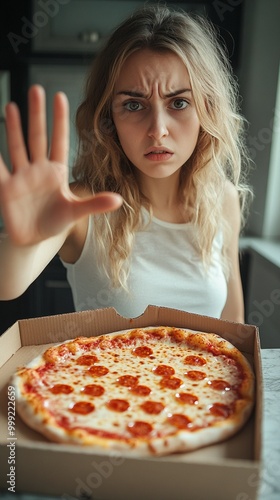 Blonde woman with serious expression gestures 'stop' with her hand while a freshly delivered pepperoni pizza sits temptingly in an open box before her, creating a conflicting scene. photo