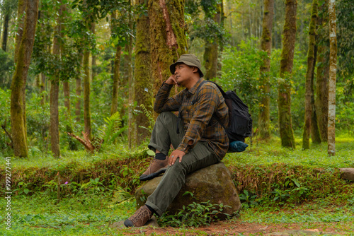 An adventurer is sitting on a large rock in a dense pine forest. An Asian man in a flannel shirt and bucket hat is sitting and resting while enjoying the tranquility of the beautiful nature.