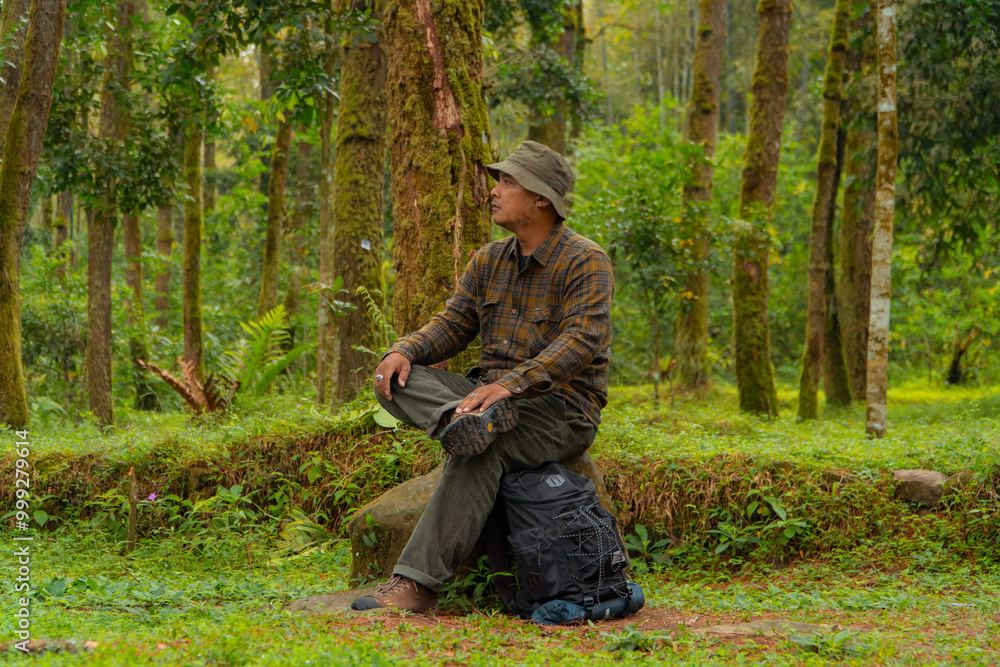 An adventurer is sitting on a large rock in a dense pine forest. An Asian man in a flannel shirt and bucket hat is sitting and resting while enjoying the tranquility of the beautiful nature. An adventurer is sitting on a large rock in a dense pine forest. An Asian man in a flannel shirt and bucket hat is sitting and resting while enjoying the tranquility of the beautiful nature.