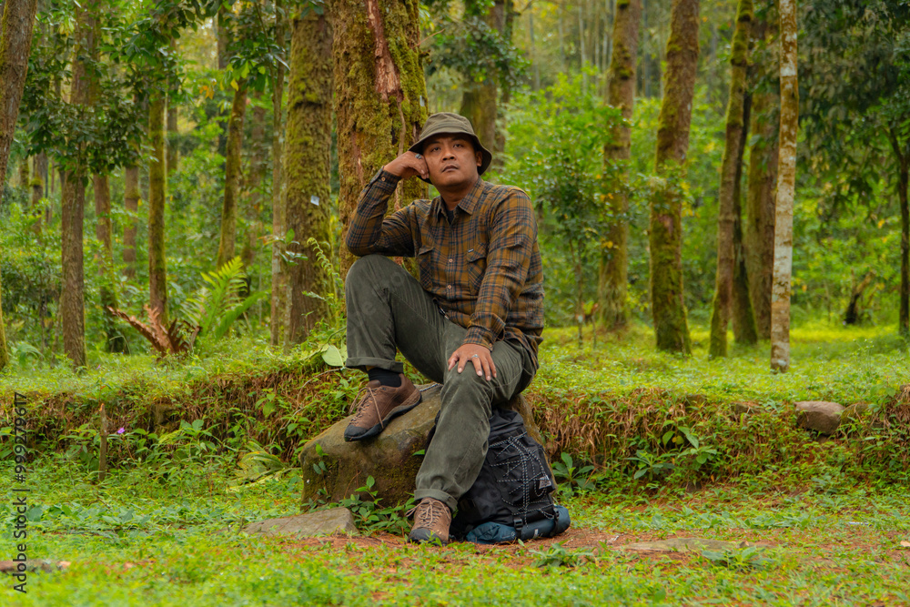 An adventurer is sitting on a large rock in a dense pine forest. An Asian man in a flannel shirt and bucket hat is sitting and resting while enjoying the tranquility of the beautiful nature. An adventurer is sitting on a large rock in a dense pine forest. An Asian man in a flannel shirt and bucket hat is sitting and resting while enjoying the tranquility of the beautiful nature.