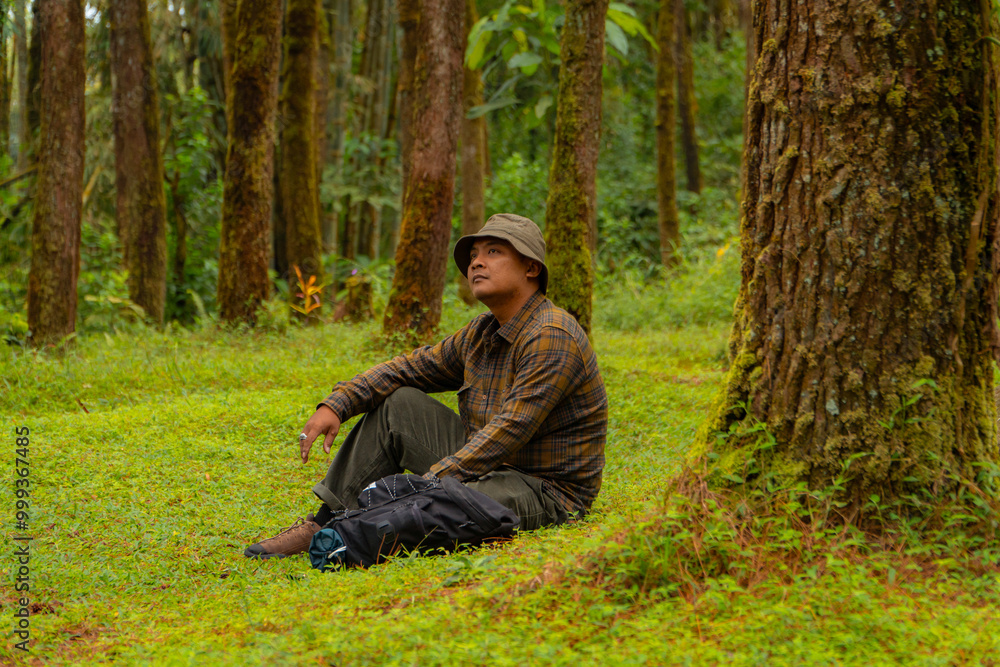 An adventurer is sitting on green grass in a lush pine forest area. Asian man in flannel shirt and bucket hat is sitting and resting while enjoying the tranquility of beautiful nature An adventurer is sitting on green grass in a lush pine forest area. Asian man in flannel shirt and bucket hat is sitting and resting while enjoying the tranquility of beautiful nature