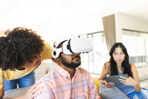 Using VR headset, man interacting with diverse friends, woman holding tablet nearby, at home photo