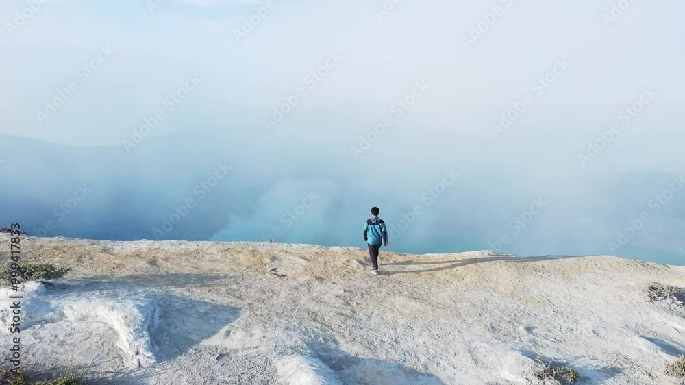 Young man walking along the top of a volcano under the rising sun with a backdrop of a turquoise sulfur lake and rocky mountains Young man walking along the top of a volcano under the rising sun with a backdrop of a turquoise sulfur lake and rocky mountains