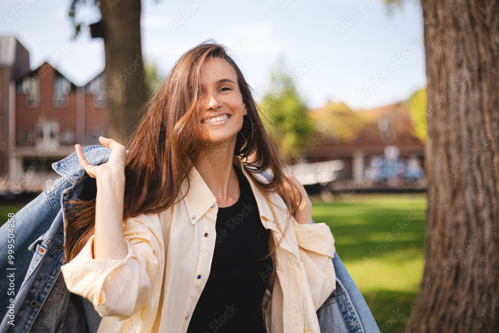 Pretty brunette woman putting on a denim jacket in the breezing air, with outdoor background. A happy woman with long dark hair, wearing jeans jacket and shirt. Girl walk in the park, look happy laugh Pretty brunette woman putting on a denim jacket in the breezing air, with outdoor background. A happy woman with long dark hair, wearing jeans jacket and shirt. Girl walk in the park, look happy laugh