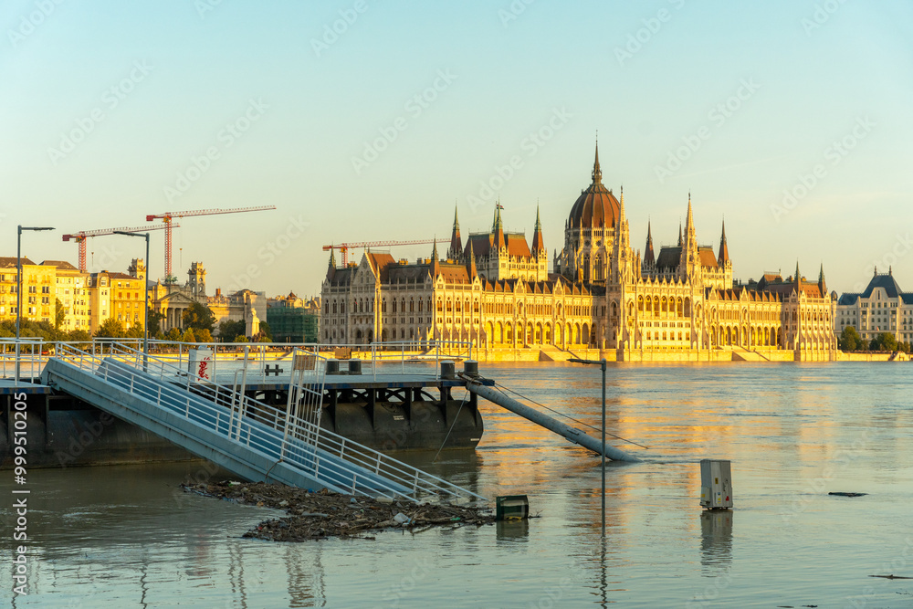 Fototapeta premium Budapest, Hungary - September 19, 2024: The parliament building in sunset golden light. Danube river overflowed its banks on the foreground.