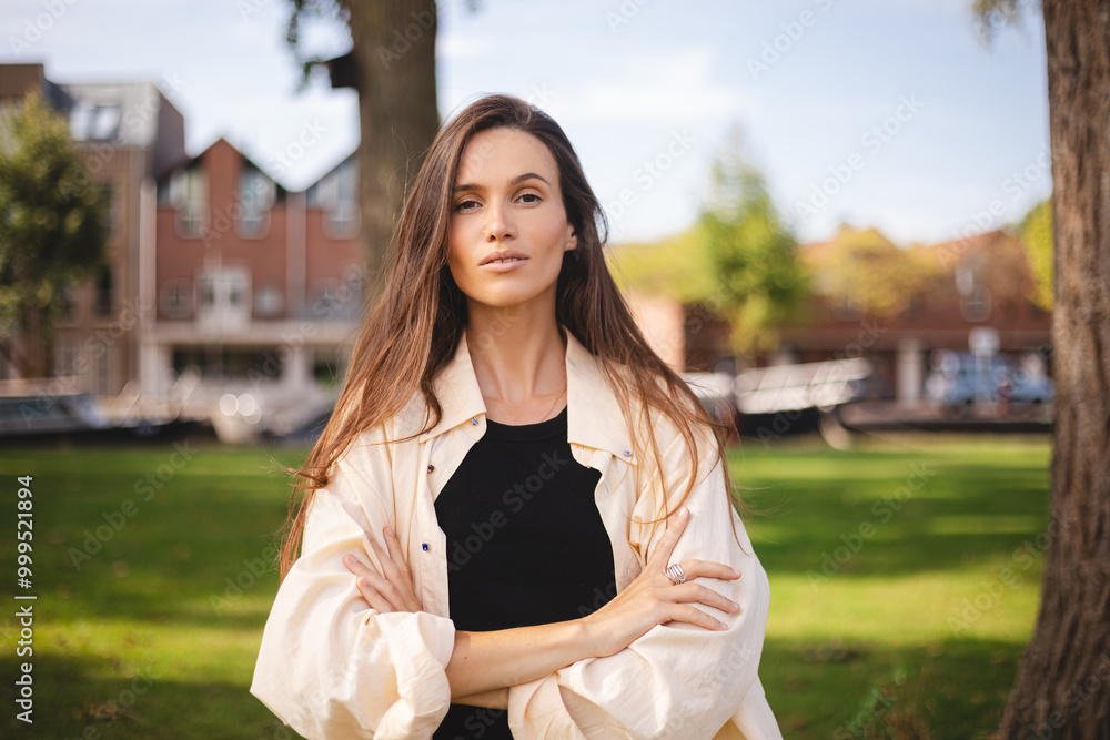 Serious brunette woman in butter color shirt standing alone in park with hands folded on chest. Successful business woman feeling confident in career development and opportunities. Serious brunette woman in butter color shirt standing alone in park with hands folded on chest. Successful business woman feeling confident in career development and opportunities.
