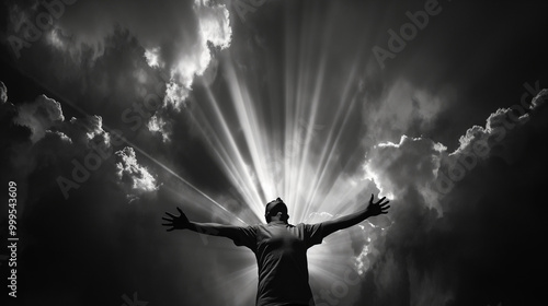 A man stands with his arms outstretched, facing the camera in black and white. He is illuminated by rays of light that emanate from behind him