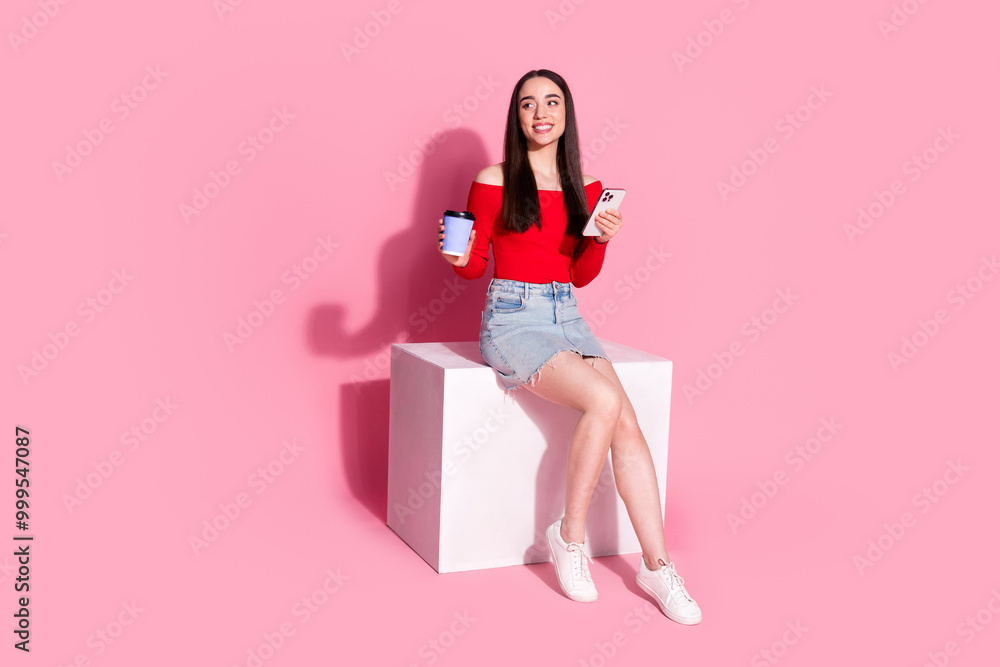 Full body portrait of pretty young girl sit cube phone coffee look empty space isolated on pink color background Full body portrait of pretty young girl sit cube phone coffee look empty space isolated on pink color background