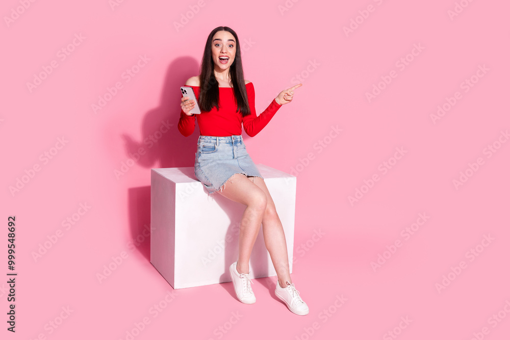 Full size photo of gorgeous young woman sit podium smart phone indicate finger empty space isolated on pink color background