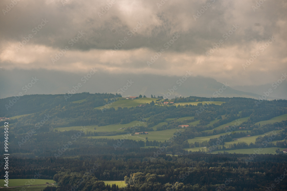 Fototapeta premium A misty, atmospheric view of rolling hills, forests, and scattered farms in Bavaria, with dark clouds covering the sky..